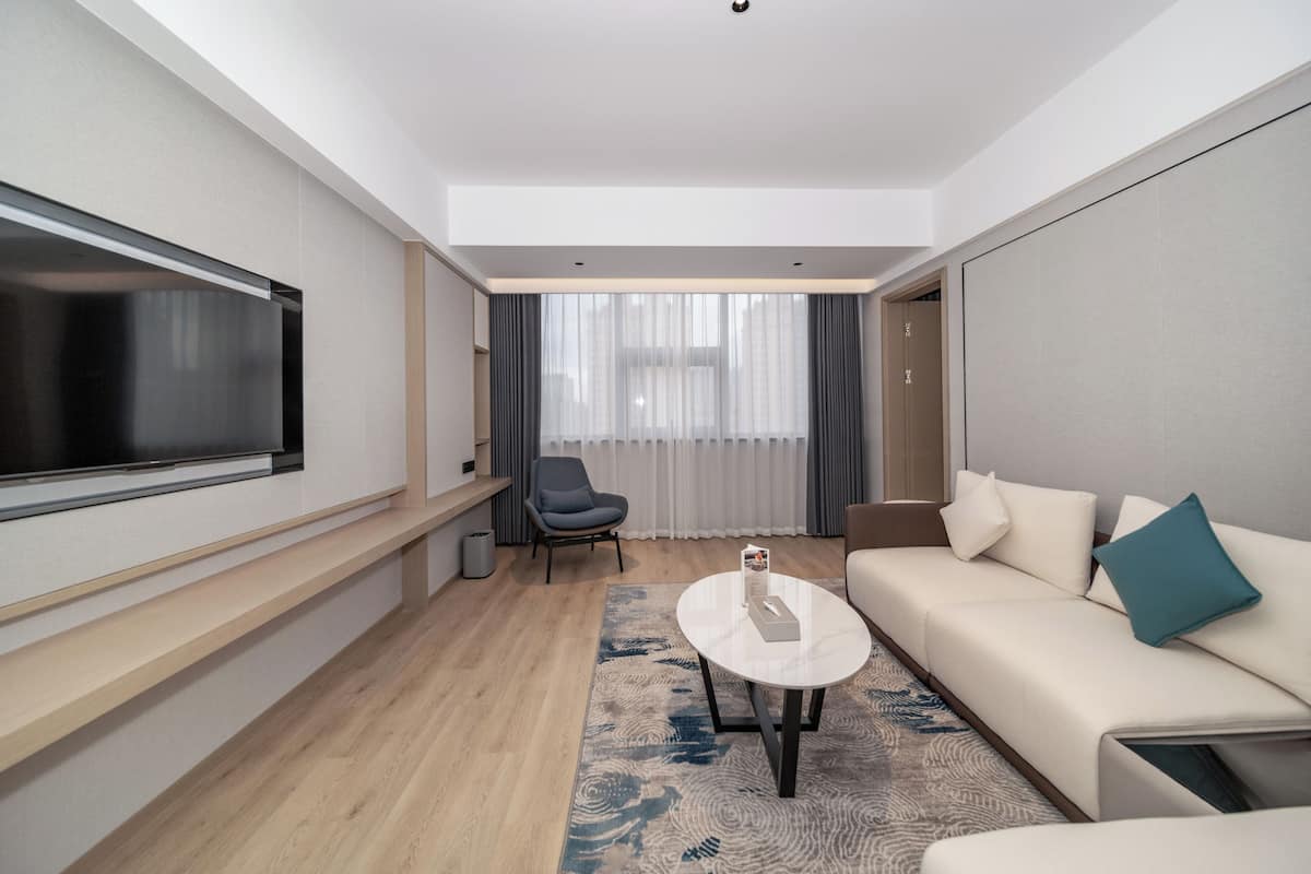 A modern hotel room with a flat-screen TV, a sofa, a chair, a coffee table, and a large window with curtains.