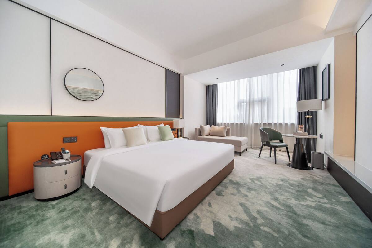 A hotel room with a large bed, a round mirror, a seating area with a chair and table, and a view of the outdoors through the window.