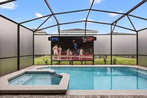 Ein Schwimmbecken mit Basketballspiel auf einer großen Leinwand, umgeben von einer überdachten Zone mit Blick auf eine Rasenfläche.