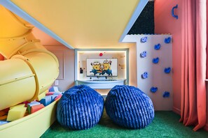 Ein Spielzimmer mit einer Rutsche, bunten Schaumstoffbausteinen und einem Fernseher, der animierte Figuren zeigt.