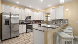 Cocina moderna con armarios blancos, electrodomésticos de acero inoxidable, encimera de granito y una zona de bar con taburetes.