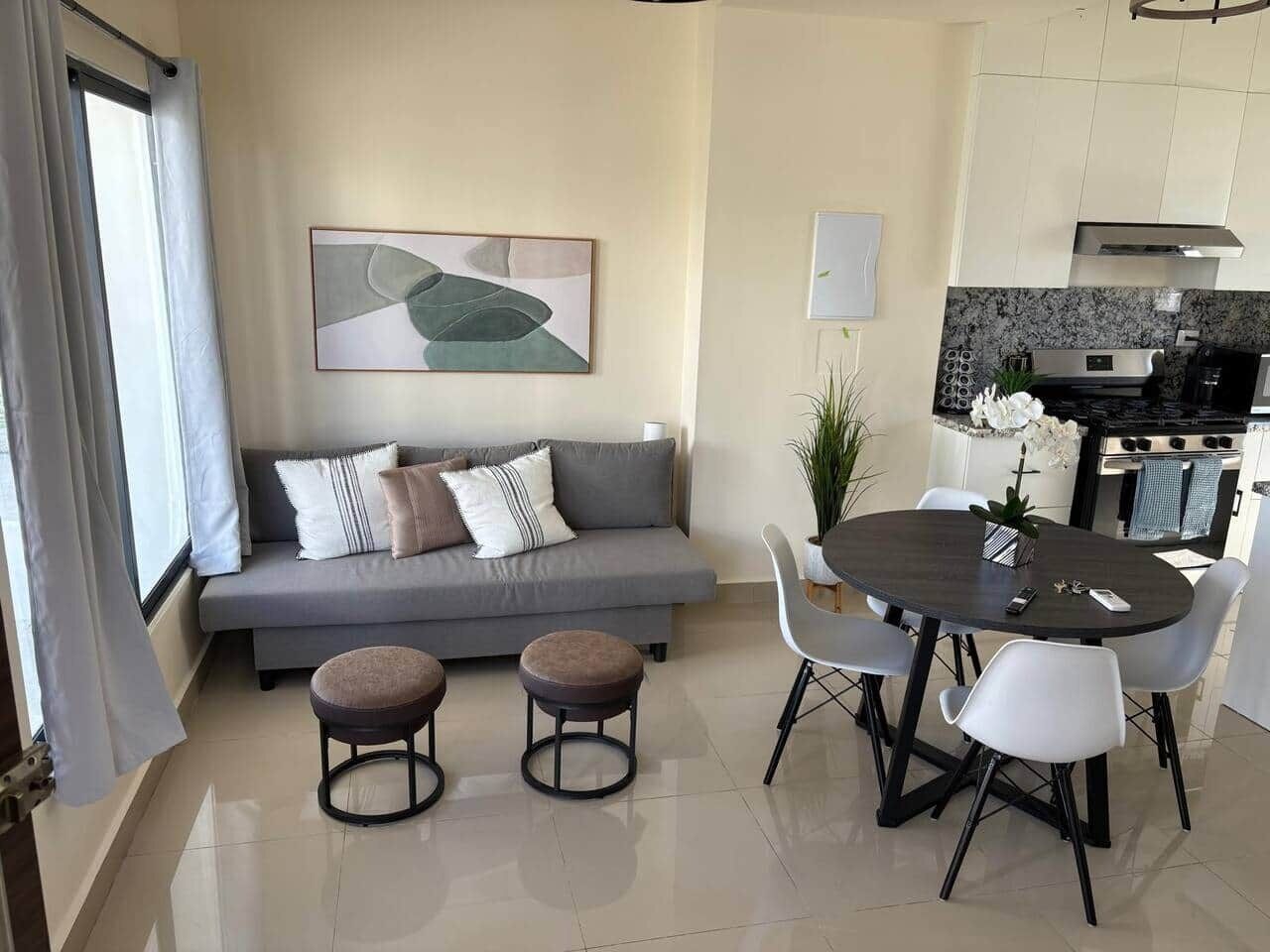 A modern living room with a grey sofa, two round ottomans, a dining table with chairs, and a kitchen area with a stove and oven.