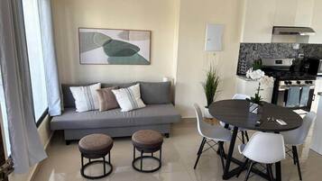 A modern living room with a grey sofa, two round ottomans, a dining table with chairs, and a kitchen area with a stove and oven.