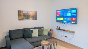 A modern living room with a grey sectional sofa, a mounted flat-screen TV displaying various apps, a framed landscape painting, and a small wooden coffee table with a plant. - Cana Life Breeze | Blue Coral Residences C1 (Punta Cana)