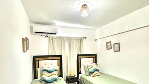 A room with two single beds, a small bedside table with a lamp, an air conditioning unit, and a ceiling light fixture. - Cana Life Breeze | Blue Coral Residences C1 (Punta Cana)
