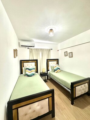 Ein Zimmer mit zwei Einzelbetten, einem kleinen Nachttisch mit Lampe, einer Klimaanlage und einer Deckenleuchte.