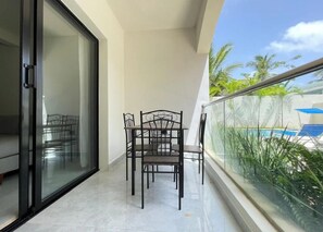 A balcony with a glass railing, a dining table with chairs, and a view of a swimming pool. - Cana Life Breeze | Los Corales Beach Residences B2 (Punta Cana)