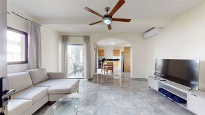 Una sala de estar moderna con un sofá, un televisor, un ventilador de techo y una cocina al fondo.