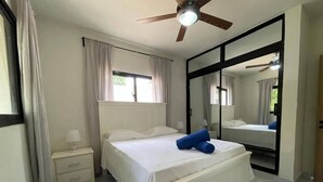 Un dormitorio con cama, mesita de noche, espejo, ventilador de techo y ventana con cortinas.