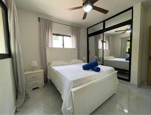 A bedroom with a bed, nightstand, mirror, ceiling fan, and window with curtains. - Cana Life Breeze | Los Corales Beach Residences B2 (Punta Cana)
