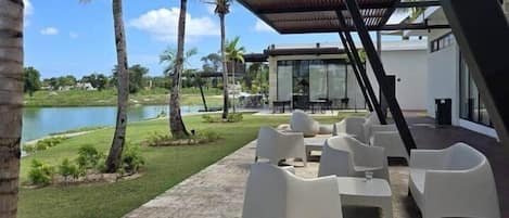 Un espacio de estar al aire libre con techo, sillas blancas modernas, una piscina y palmeras al fondo.