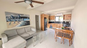 A modern living room with a grey sofa, a glass coffee table, and a dining area with wooden chairs and a table. - Cana Life Breeze | Los Corales Beach Residences B4 (Punta Cana)
