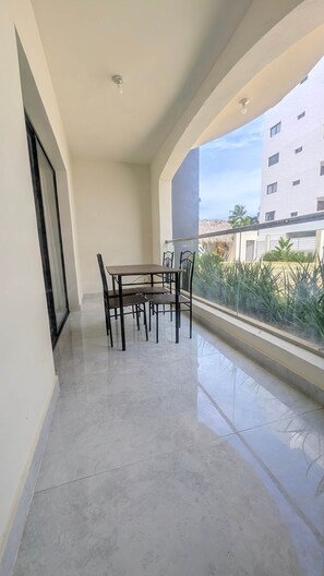 A balcony with a dining table and chairs, a glass railing, and a view of a building and greenery. - Cana Life Breeze | Los Corales Beach Residences B1 (Punta Cana)