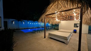 Ein überdachter Außenbereich mit Strohdach, Pool, Liegestühlen und einer Liege.