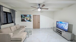 A living room with a sofa, TV, ceiling fan, and a door. - Cana Life Breeze | Los Corales Beach Residences B3 (Punta Cana)