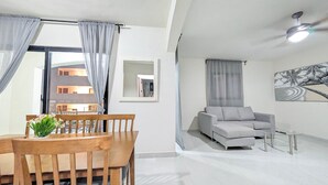 Una sala de estar moderna con un sofá gris, una mesa de comedor con sillas, un ventilador de techo y un cuadro en la pared.
