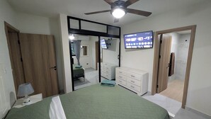 Un dormitorio con cama, armario, espejo, ventilador de techo y televisor que muestra una plataforma de streaming.
