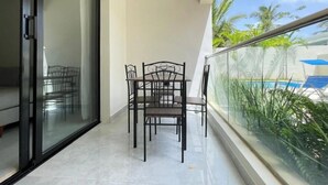 Un balcón con barandilla de vidrio, una mesa de comedor con sillas y vista a una piscina.