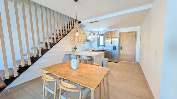 A modern kitchen with a central island, wooden staircase, and a dining area with a table and chairs. - Cana Life Luxury | Azure Sky Penthouse (Punta Cana)
