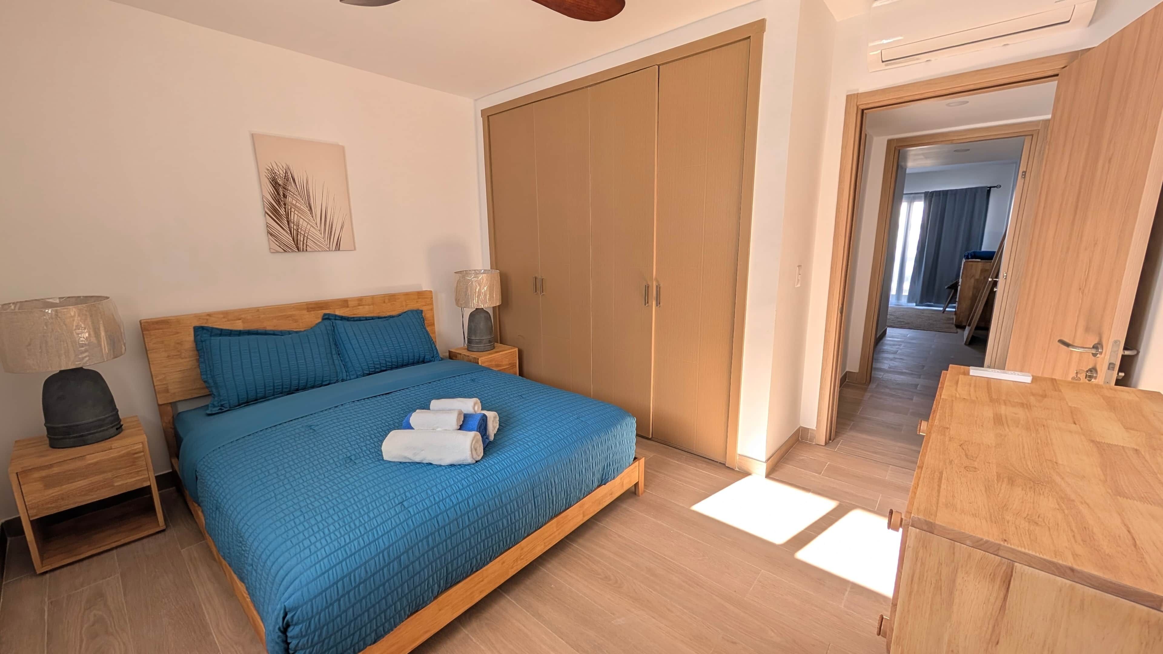 A bedroom with a wooden bed, a nightstand with a lamp, a wardrobe, and a view into another room.