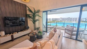 Una sala de estar moderna con un televisor de pantalla plana grande, un sofá y un comedor con vistas a una piscina y edificios de hotel.