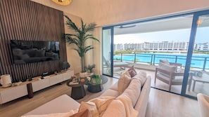 Ein modernes Wohnzimmer mit großem Flachbildfernseher, einer Couch und einem Essbereich mit Blick auf einen Pool und Hotelgebäude.