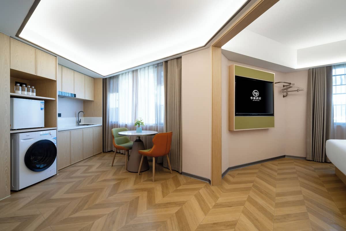 A modern hotel room with a kitchen area, a dining table with chairs, a TV mounted on the wall, and a large window with curtains.