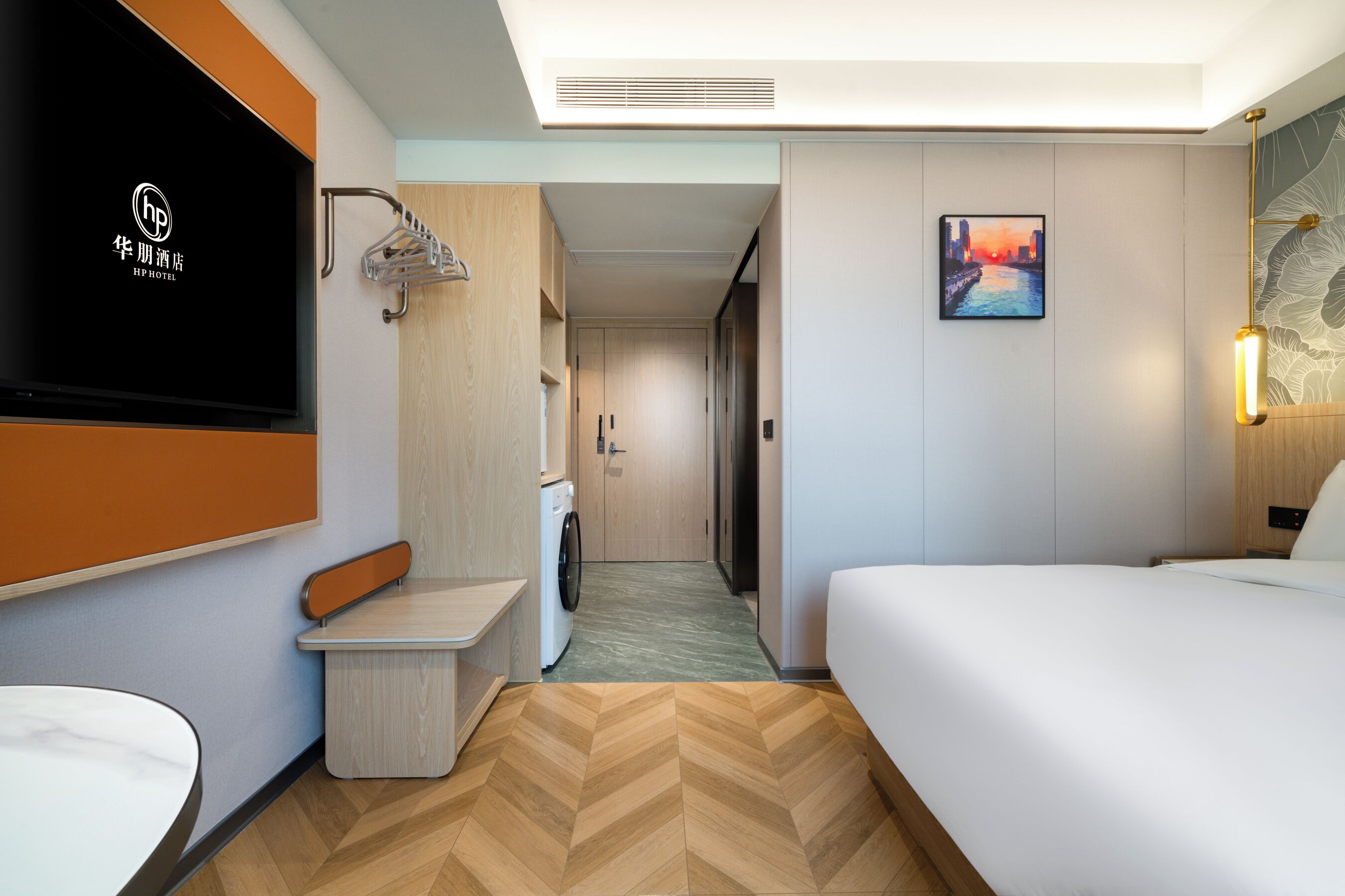 A hotel room with a bed, a small table, a washing machine, a TV, and a painting on the wall.