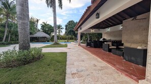 A covered outdoor seating area with a pool, palm trees, and a thatched-roof structure. - Cana Life Luxury | Villa Cueva (Punta Cana)