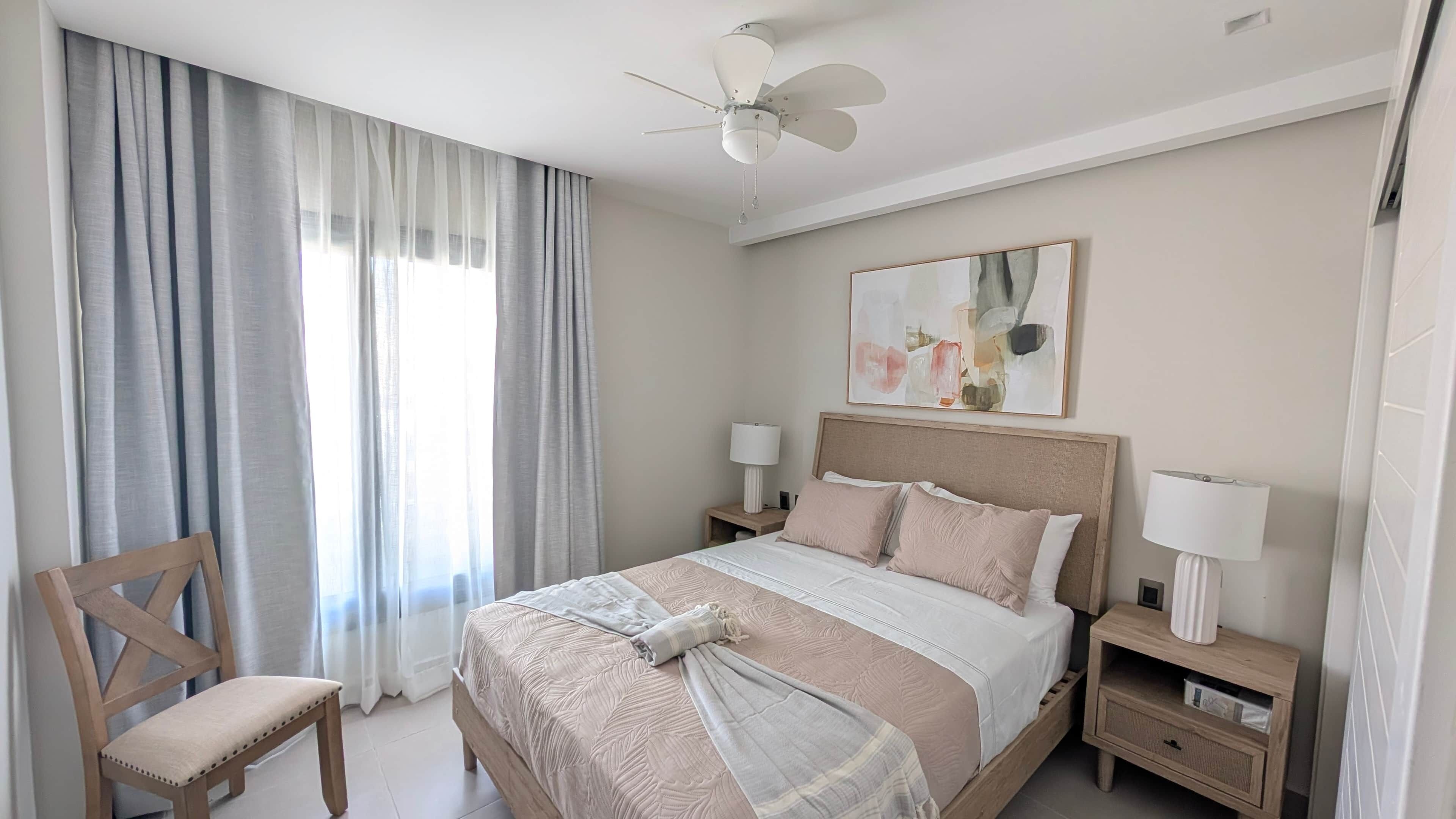 Un dormitorio con una cama, una silla, un ventilador de techo, una mesita de noche con lámpara y un cuadro en la pared.