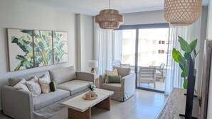 Uma sala de estar moderna com sofá, mesa de centro e área de jantar com cadeiras. Há uma janela grande com cortinas, um lustre e um vaso de planta.