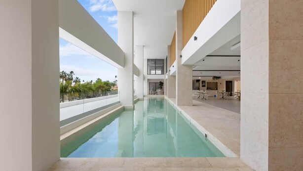 Piscina interior moderna com água cristalina, cercada por paredes e colunas brancas, e vista para palmeiras lá fora.