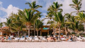 A beach resort with palm trees, lounge chairs, and a building with a thatched roof. - Cana Life Luxury | Sapphire Sky Pool Penthouse (Punta Cana)