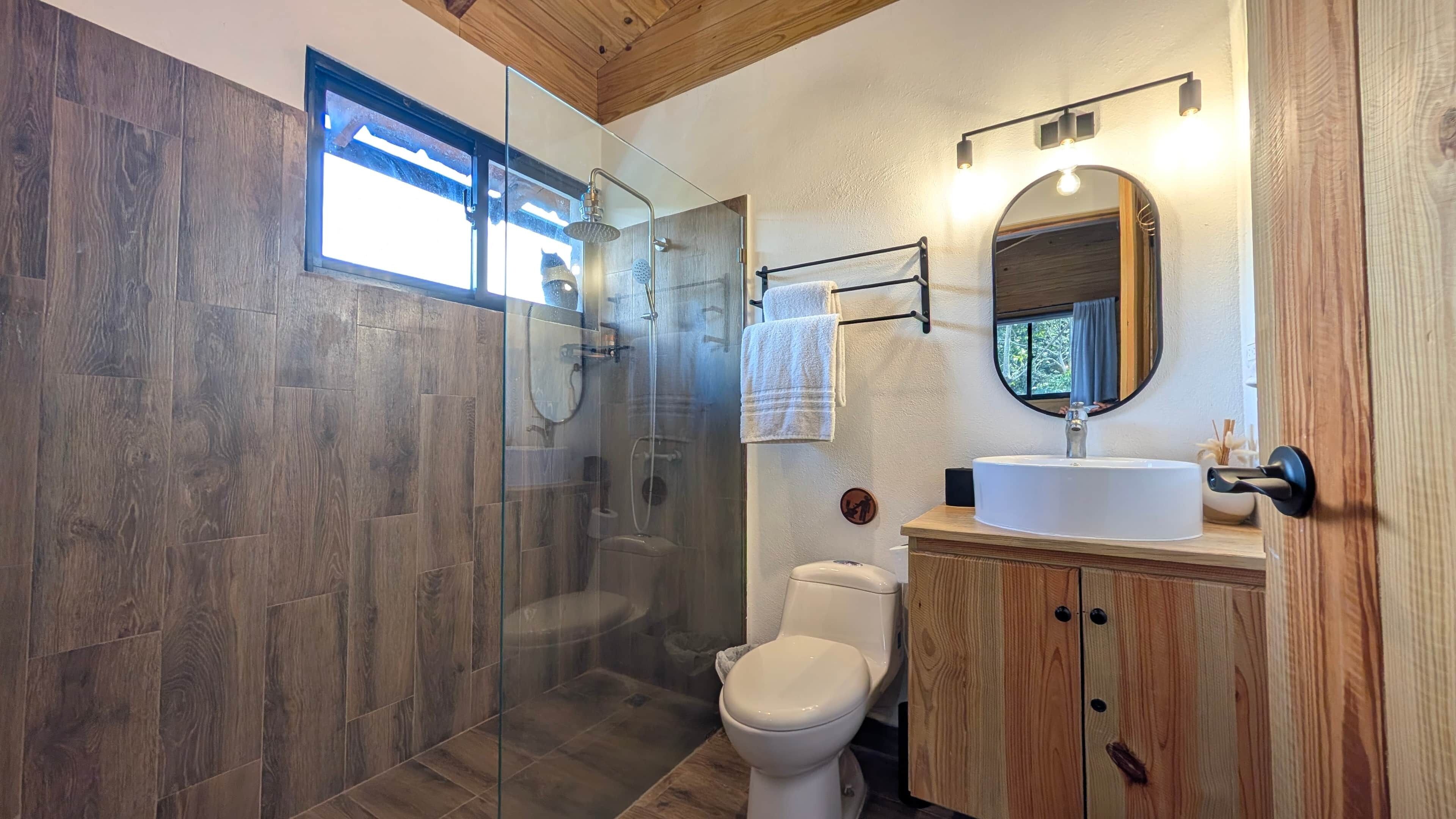 Une salle de bain moderne avec une douche à parois de verre, des toilettes blanches et un meuble-lavabo en bois surmonté d’un miroir rond.