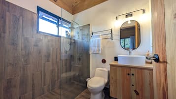 Une salle de bain moderne avec une douche à parois de verre, des toilettes blanches et un meuble-lavabo en bois surmonté d’un miroir rond.