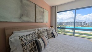 Een hotelkamer met een groot bed, uitzicht op een zwembad en een complex van gebouwen op de achtergrond.