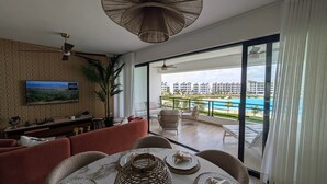 Een moderne woonkamer met een grote televisie, een eethoek met een tafel voor twee personen, en een balkon met uitzicht op een zwembad.