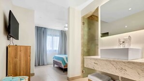 Habitación de hotel con armario de madera, televisor, cama con cubrecama azul y baño con lavamanos y toallero de mármol.