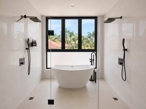 A modern bathroom with a freestanding bathtub, two showerheads, and a large window with a view of palm trees. - Cana Life Reserve | Villa Azure (Punta Cana)
