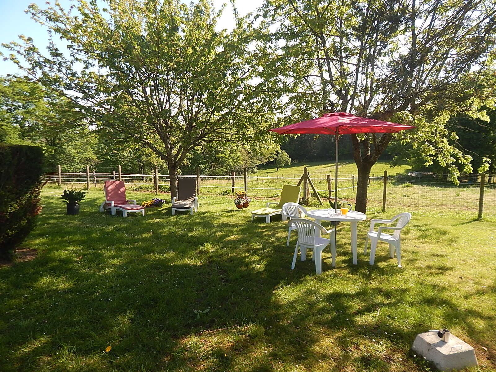 Ein grünes Areal im Freien mit einem roten Sonnenschirm, weißen Stühlen und einem Tisch.