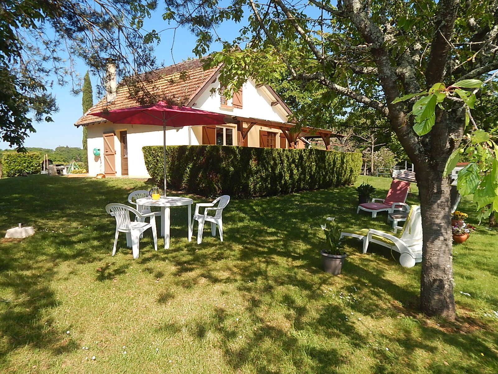 A house with a red roof, a white exterior, and a well-maintained lawn. There is a table with chairs and a red umbrella.