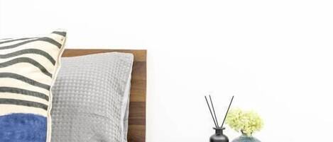 Una cama bien tendida con almohadas, una mesita de noche con un florero y difusor de aroma, y un cuadro en la pared.