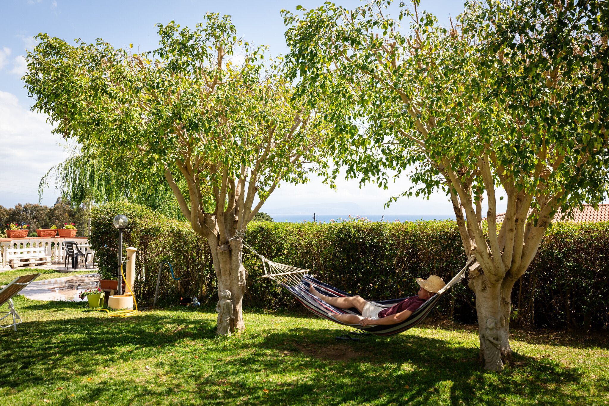 A hammock between two trees in a grassy area with a building and a view of the sea in the background.
