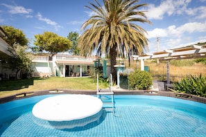 Ein Schwimmbecken mit Whirlpool, umgeben von einer Terrasse mit einer Pergola, Palmen und einem Haus im Hintergrund.