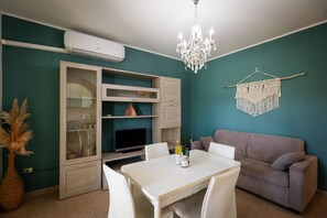 Une pièce aux murs de couleur turquoise, avec une table à manger blanche, un canapé gris, un lustre, un téléviseur et un élément décoratif mural.