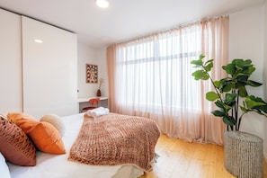 Ein Schlafzimmer mit einem Bett, Kissen, einem Nachttisch, einer Pflanze und einem Fenster mit Vorhängen.