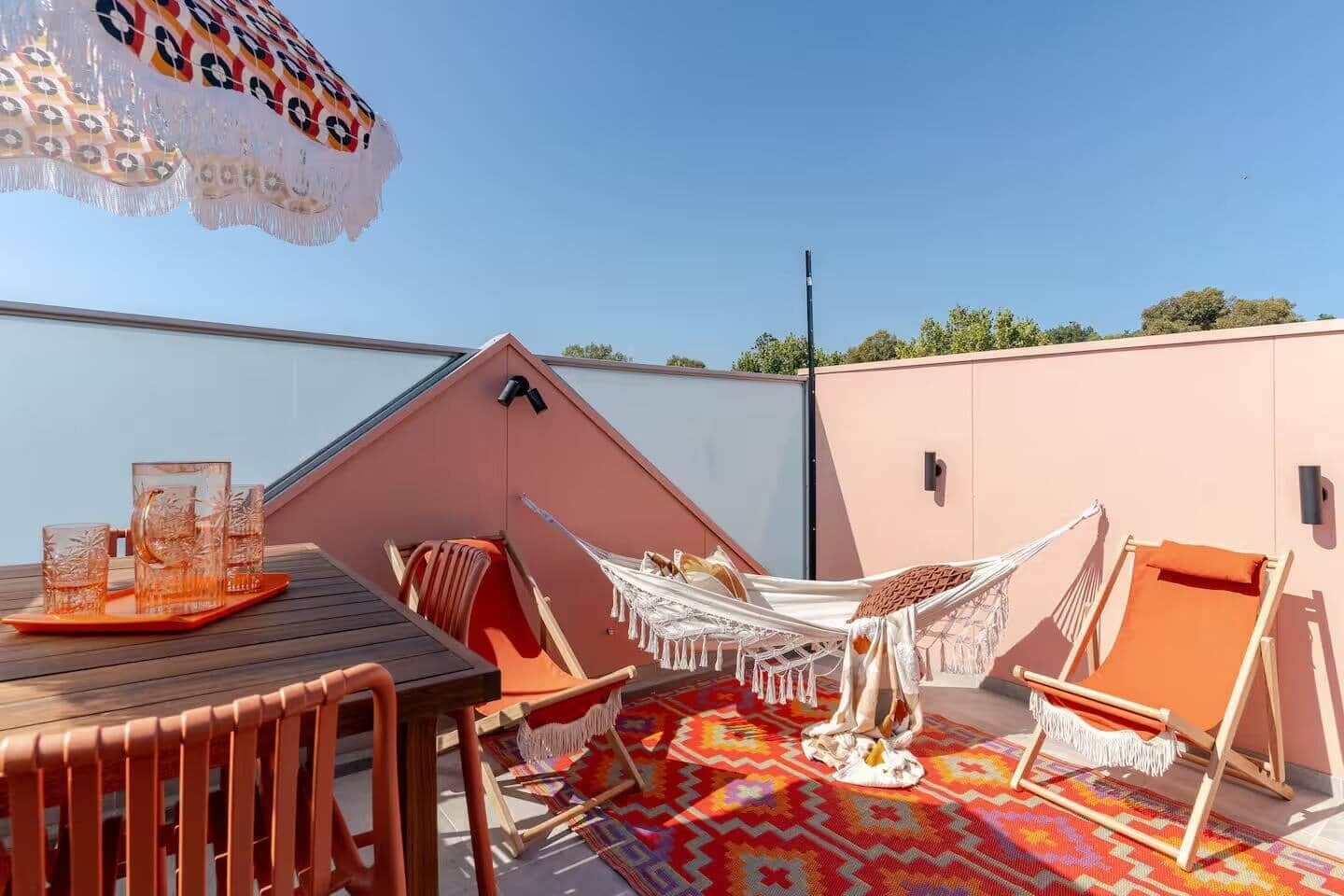 A rooftop terrace with a wooden table, two hammocks, a colorful rug, and a clear blue sky.