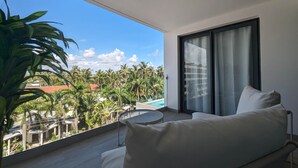Een balkon met uitzicht op palmbomen en een zwembad, een witte bank en een ronde tafel.