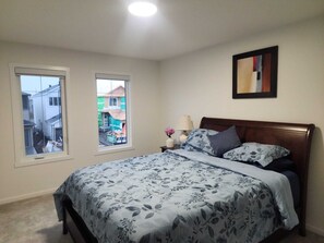 Ein Schlafzimmer mit einem Bett, zwei Fenstern, einem Nachttisch, einer Lampe und einem gerahmten Bild an der Wand.