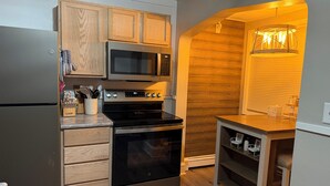 A compact kitchen with wooden cabinets, a stainless steel refrigerator, a microwave, an oven, and a small wooden table with a hanging light fixture.